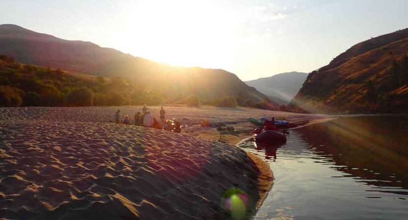 a watercraft rests on a calm river while the sun sets over mountains in the background